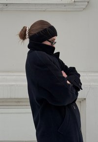 Manteau en laine noire à col montant, associé à un bandeau noir et un chouchou de couleur claire. Le sujet est les bras croisés, de profil.