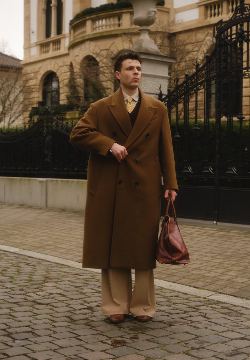 Cappotto doppiopetto marrone, pantaloni beige a gamba larga, scarpe di pelle marroni e una borsa marrone, su uno sfondo di pietra e ferro battuto.