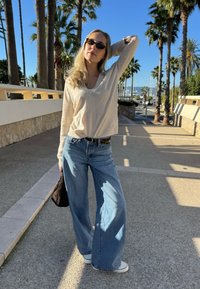 Haut beige clair à manches longues, jean large bleu ample et baskets blanches. Tient un petit sac sombre le long d'une promenade ensoleillée bordée de palmiers.