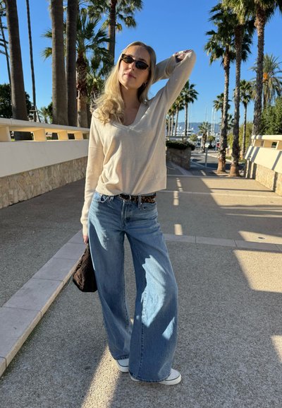 Top de manga larga beige claro, vaqueros azules de pierna ancha y corte suelto, y zapatillas blancas. Sostiene una pequeña bolsa oscura junto a un paseo soleado con palmeras.