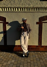 Homem em um blazer xadrez sobre um suéter de gola alta castanho, calças claras e sapatos pretos e brancos, encostado a uma parede texturizada com sombras.