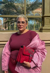 Borsa a tracolla rossa con dettagli decorativi in argento, tenuta contro una giacca di raso rosa. Sullo sfondo si possono vedere foglie di palma e dettagli architettonici.