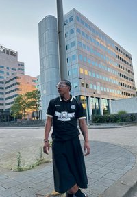 Μαύρο πουκάμισο με λευκές και κίτρινες λεπτομέρειες, με γραφικό σχέδιο. Συνδυάζεται με μακριά μαύρη φούστα. Αστική τοποθεσία με μοντέρνα κτίρια.