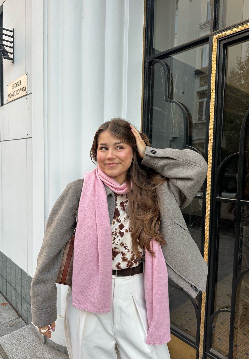 Cappotto grigio oversize, sciarpa rosa chiaro, top con motivi e pantaloni crema. Capelli lunghi e mossi e borsa marrone. Elementi decorativi dell'edificio.