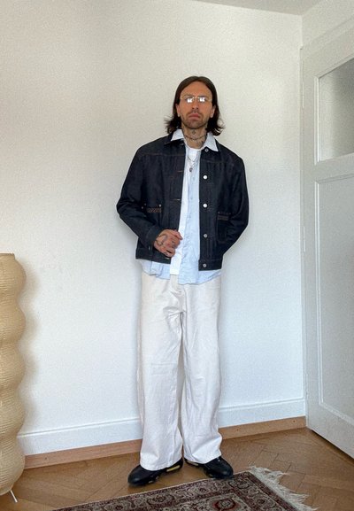 Hombre con cabello hasta los hombros y tatuajes, de pie en interiores, lleva gafas, chaqueta de mezclilla negra, camisa azul clara, pantalones blancos anchos y zapatos negros.