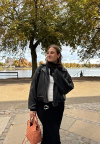 Czarna skórzana kurtka o cropped fasonie na białym topie, w zestawieniu z czarną spódnicą. Trzyma plecioną pomarańczową torb�ę o teksturowanym wykończeniu.