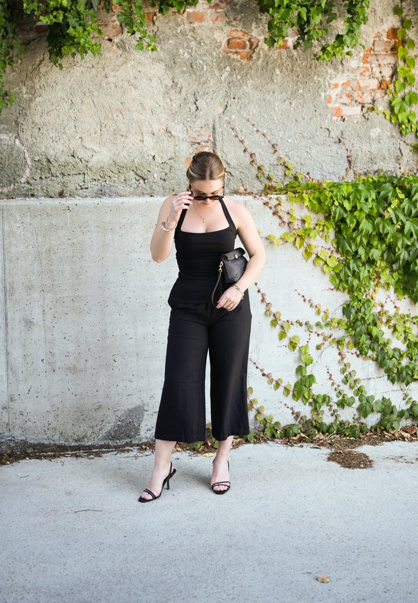 Donna con tuta nera senza maniche che sistema gli occhiali da sole, tiene una borsa nera, in piedi accanto a un muro di cemento e mattoni coperto di edera.
