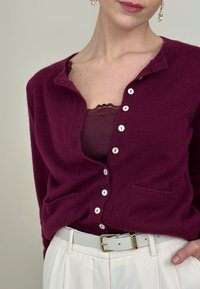 Cardigan bordeaux avec un col en V, tissu texturé, et boutons blancs. Porté sur un débardeur noir bordé de dentelle, assorti à un pantalon blanc et une ceinture.
