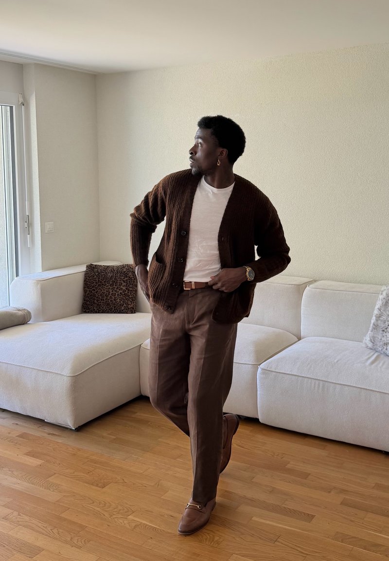 Homme en cardigan marron, chemise blanche et pantalon marron debout sur un sol en bois près d'un canapé d'angle blanc avec des coussins léopard et gris.