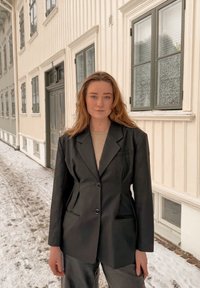 Γυναίκα με μακριά μαλλιά, που φοράει μαύρο σακάκι και μπεζ τοπ, στέκεται σε εξωτερικό χώρο σε χιονισμένο δρόμο μπροστά από ένα κρεμ ξύλινο κτήριο.