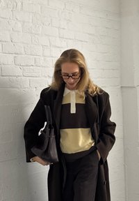 Hnedý kabát, krémový polo s tmavohnedými a žltými pruhmi, čierne nohavice, drží čiernu tašku. Svetlá textúra na bielom tehlovom pozadí.