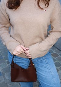 Femme en pull beige et jeans bleu tenant un petit sac à main en cuir marron avec des bagues aux doigts, sur un fond de pavés.