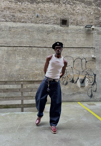 Persona vestida con una camiseta sin mangas blanca, boina negra con el logo de NY, pantalones vaqueros amplios de color azul oscuro y zapatos marrones con cordones. Lleva un collar de cuentas y un colgante.