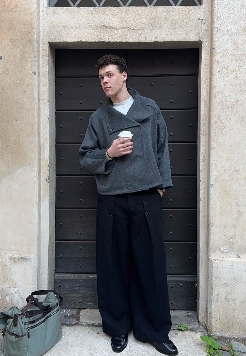 Jeune homme portant un pantalon large noir et une veste grise, tenant une tasse de café, appuyé contre une porte en bois sombre encadrée de murs en pierre. Sac posé au sol à proximité.