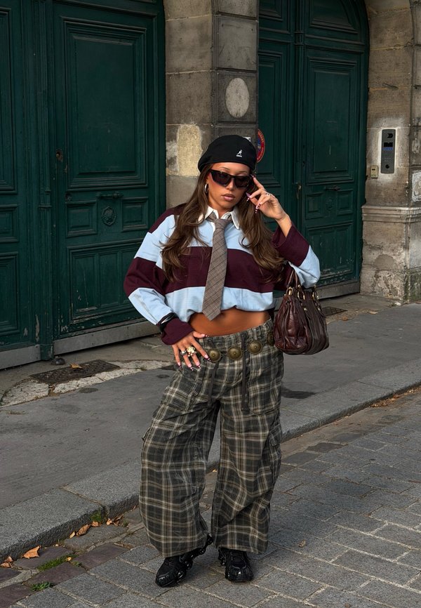 Jeune femme portant un maillot de rugby court, une cravate, un pantalon large à carreaux, un béret noir, des lunettes de soleil et tenant un sac à main marron dans une rue de la ville.