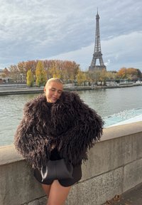 Jeune femme portant un grand manteau en fourrure sombre, appuyée sur un mur au bord de la rivière, avec des arbres d'automne et la Tour Eiffel en arrière-plan sous un ciel nuageux.