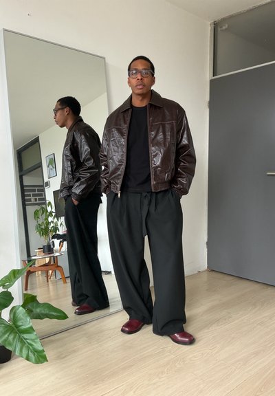 Hombre con chaqueta de cuero marrón, pantalones anchos negros y zapatos rojos, de pie con las manos en los bolsillos, reflejado en un espejo alto en una habitación minimalista.