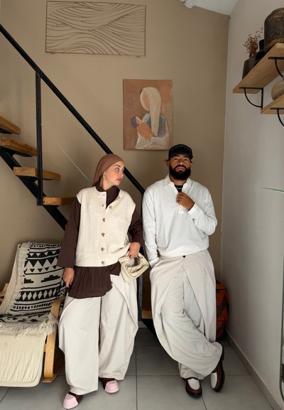 Dos personas están de pie en el interior cerca de unas escaleras; una lleva un vestido marrón, chaleco crema y pañuelo en la cabeza sosteniendo una bolsa, la otra lleva una blusa blanca, gorra y pantalones anchos crema.