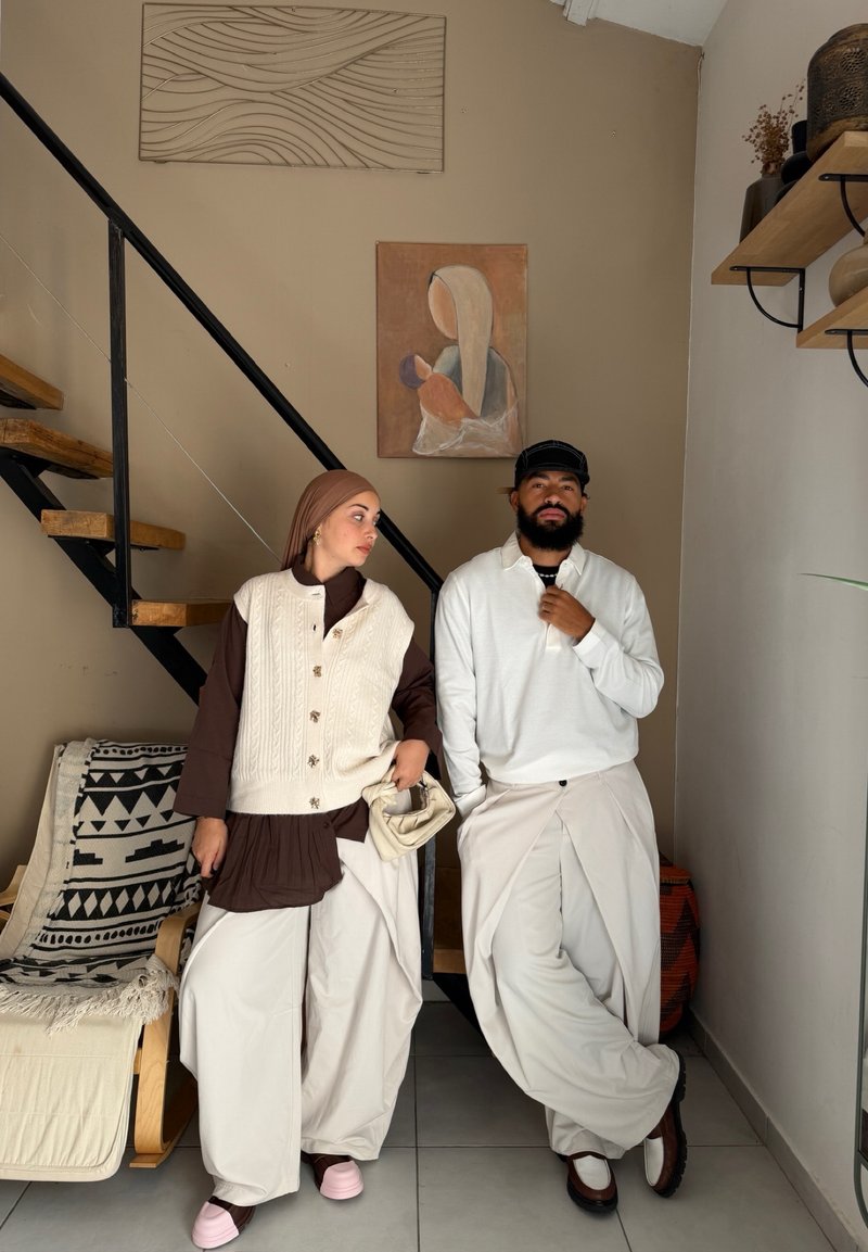 Dos personas están de pie en el interior cerca de unas escaleras; una lleva un vestido marrón, chaleco crema y pañuelo en la cabeza sosteniendo una bolsa, la otra lleva una blusa blanca, gorra y pantalones anchos crema.