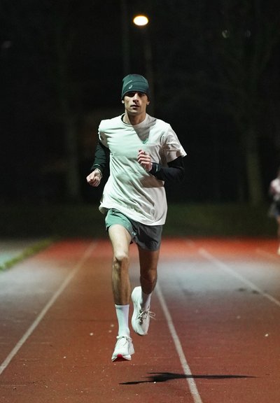 Corredor masculino con gorro y pantalones cortos, corriendo en una pista al aire libre por la noche bajo luz artificial.