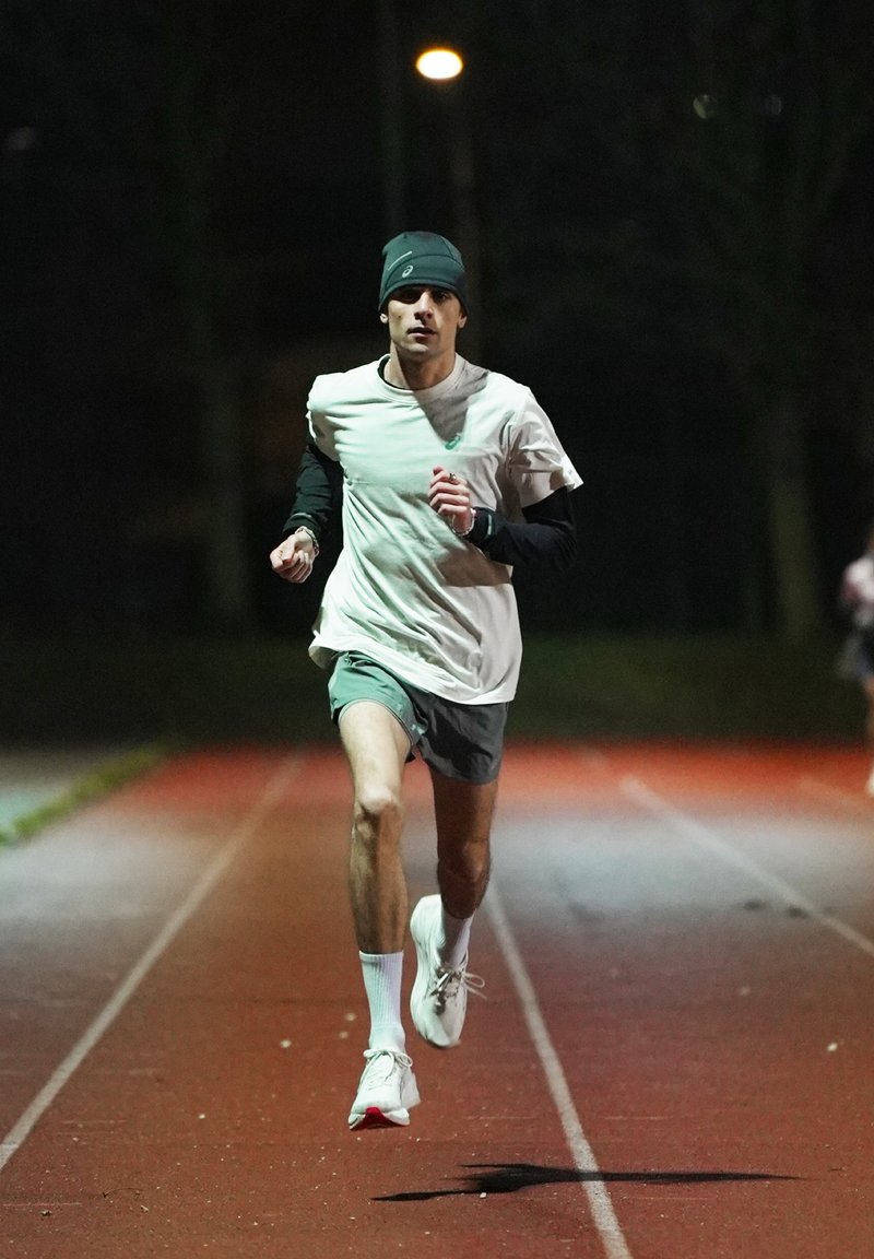 Άνδρας δρομέας που φοράει σκουφάκι και βερμούδα, τρέχει σε εξωτερικό στίβο τη νύχτα υπό τεχνητό φωτισμό.