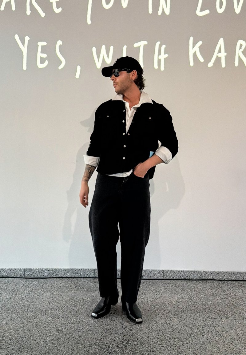Man wearing black cap, sunglasses, black jacket, white shirt, and black pants stands against white wall with partial handwritten text.