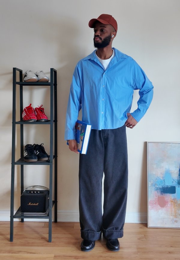 Ein Mann mit roter Kappe und blauem Hemd hält ein Buch, steht neben einem Schuhregal mit Turnschuhen und einem Lautsprecher, in der Nähe eines abstrakten Gemäldes.