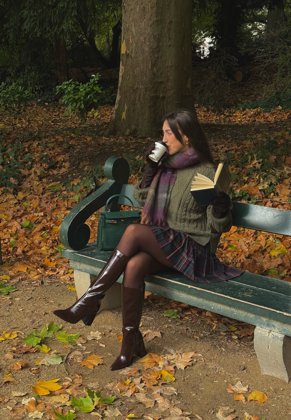 Strikket olivengrønn genser, rutete skjørt med nyanser av marineblått og burgunder, brune skinnstøvler opp til knærne, holder en bok og en kaffekopp.