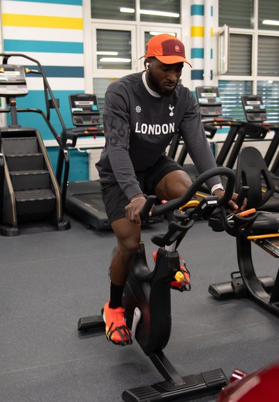 Hombre con gorra naranja y zapatos brillantes pedaleando en bicicleta estática en un gimnasio con cintas de correr al fondo.