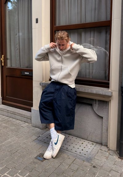 Joven ajustándose la sudadera con capucha, de pie en una acera de la ciudad cerca de una ventana y una puerta, lleva pantalones cortos anchos, calcetines blancos y zapatillas Nike blancas.