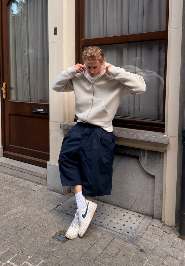 Ung mand justerer sin hættetrøje, står på en byfortov nær et vindue og en dør, iført brede shorts, hvide sokker og hvide Nike-sneakers.