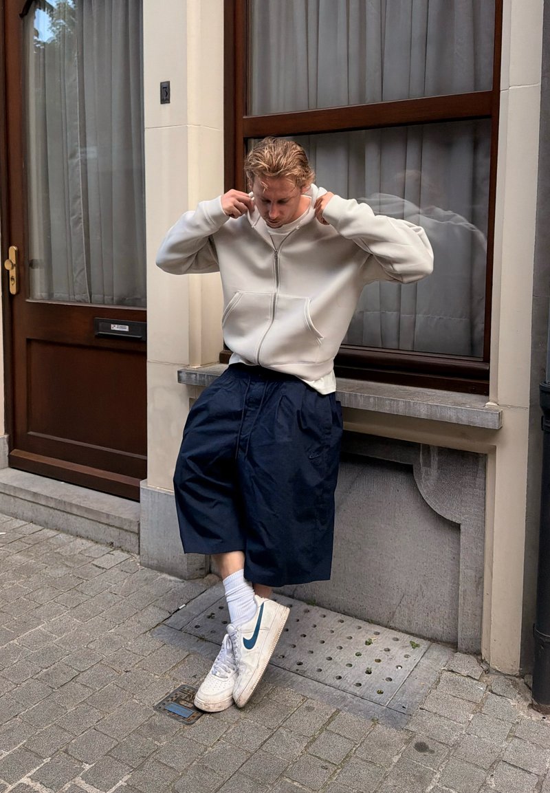Jeune homme ajustant son sweat à capuche debout sur un trottoir en ville près d'une fenêtre et d'une porte, portant un short large, des chaussettes blanches et des baskets blanches Nike.