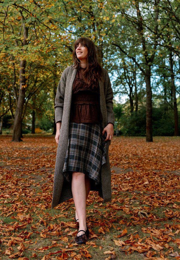 Une femme en long manteau gris et en jupe à carreaux se promène dans un parc automnal couvert de feuilles mortes.
