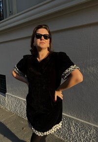 Zwarte fluwelen jurk met pofmouwen en een witte stippenrand. Model draagt een zonnebril en staat met de handen in de zij tegen een gestructureerde muur.