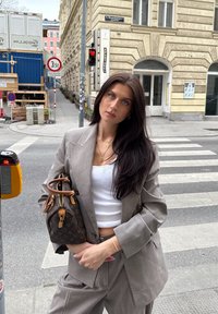 Costum gri croit cu un sacou structurat și pantaloni largi. Top alb dedesubt, ținând o geantă de mână cu un model maro. Cadru urban.