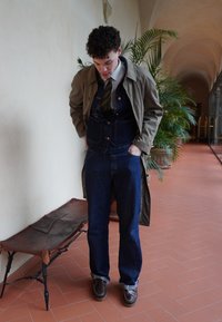 Ένα άτομο σε τζιν σαλοπέτα, καπιτονέ παλτό και καφέ παπούτσια στέκεται μέσα σε κλειστό χώρο. Σημαντικές λεπτομέρειες περιλαμβάνουν μια ριγέ γραβάτα και ύφασμα σε σκούρο μπλε χρώμα.