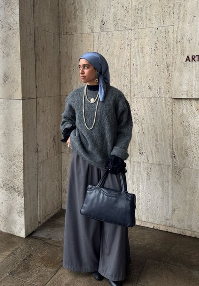 Mujer con un suéter gris, pantalones anchos grises, un pañuelo azul en la cabeza, collares de perlas y sosteniendo un bolso negro frente a un fondo de pared de piedra.