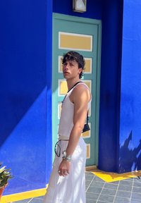 Top corto blanco, falda blanca, accesorios de metal y bolsa negra pequeña frente a una vibrante pared azul y una puerta verde claro con acentos amarillos.