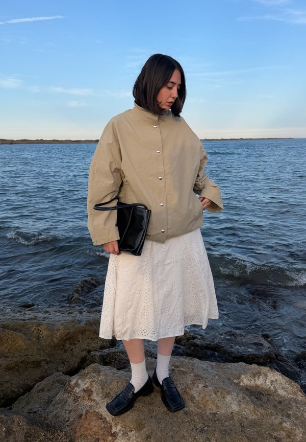 Femme portant une veste beige, une jupe blanche, des chaussettes blanches et des chaussures noires, tenant un sac noir, debout sur des rochers au bord de la mer sous un ciel clair.