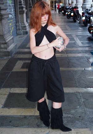 Top corto negro con cuello halter y cortes, pantalones cortos negros con cordón, y botas de flecos hasta la rodilla. Modelo sosteniendo un objeto plateado, de pie sobre pavimento de piedra.