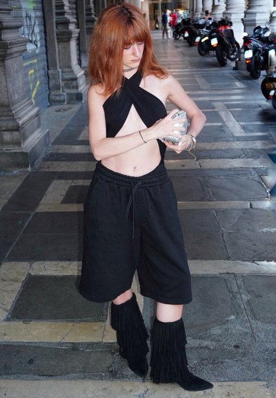 Top corto negro con cuello halter y cortes, pantalones cortos negros con cordón, y botas de flecos hasta la rodilla. Modelo sosteniendo un objeto plateado, de pie sobre pavimento de piedra.