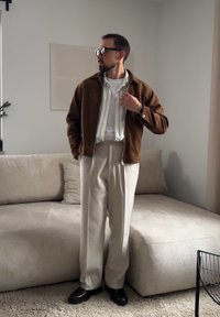 Homme portant une veste marron, une chemise rayée, un pantalon beige et des chaussures noires, debout à côté d'un canapé beige dans un salon minimaliste.