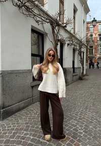 Jeune femme aux longs cheveux blonds portant un pull blanc, un pantalon marron foncé, des chaussures beiges et des lunettes de soleil, debout dans une rue pavée.