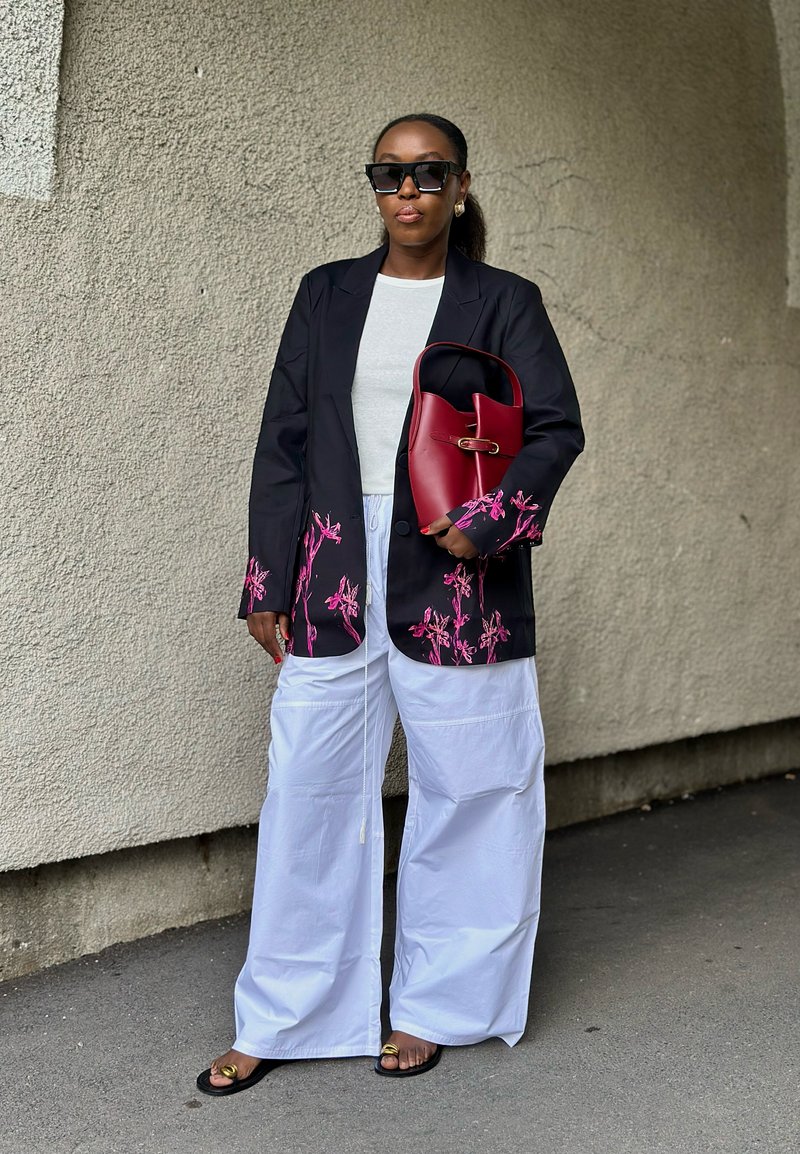 Nero blazer con ricami floreali rosa, t-shirt bianca, pantaloni bianchi oversize e una borsa rossa. Gli occhiali da sole neri e i sandali piatti completano il look.
