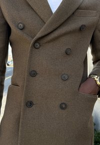 Καφέ διπλόκοντο παλτό με μαλακό, υφασματώδη υλικό. Διαθέτει έξι μαύρα κουμπιά και τσέπη στο στήθος. Φοράει μεταλλικό ρολόι.
