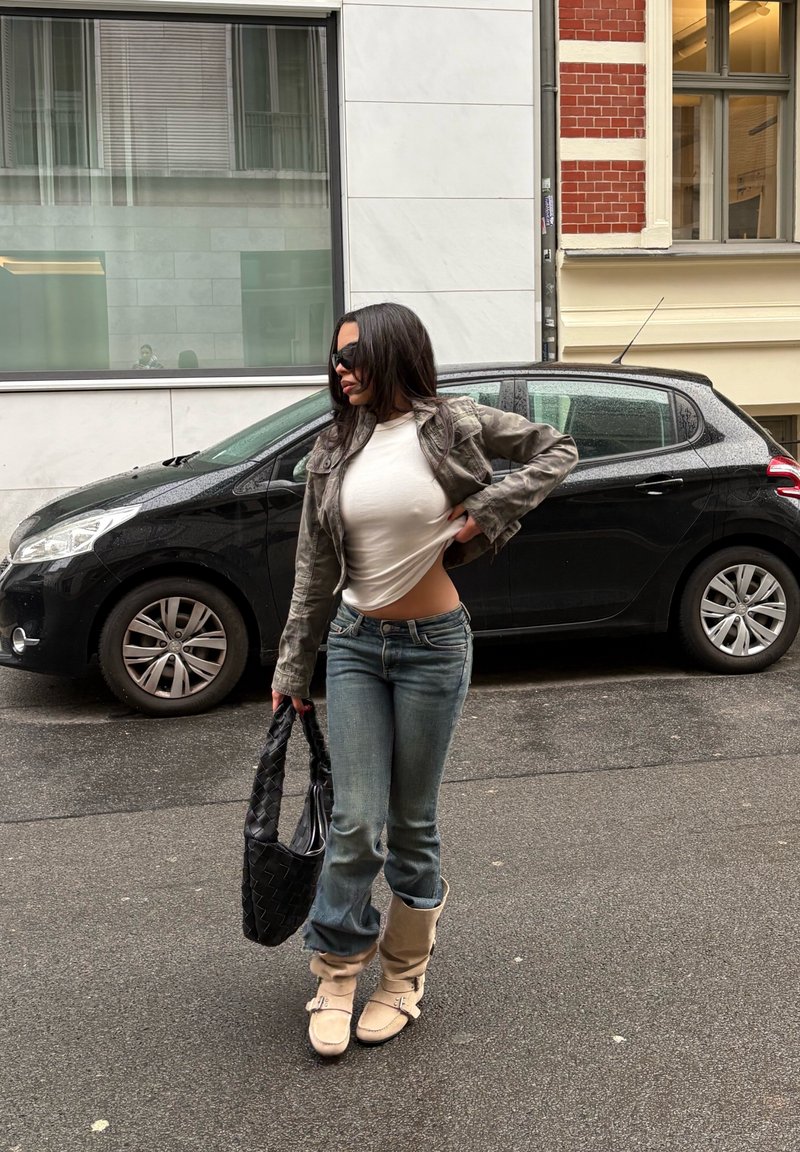 Sac à main noir tissé, veste en cuir grise raccourcie, t-shirt blanc, jean bleu, bottes beiges ; rue urbaine, voitures en arrière-plan.