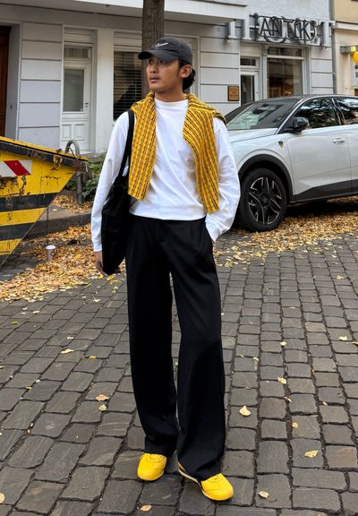 Pantalones anchos negros, camisa blanca de manga larga y un chaleco amarillo texturizado. Zapatos amarillos con un diseño deportivo. El fondo incluye una escena callejera.