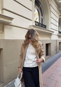 Béžový vlněný kabát přes bílý svetr s copánkovým vzorem, doplněný černými kalhotami. Drží bílou kabelku; vlasy upravené do volných vln.