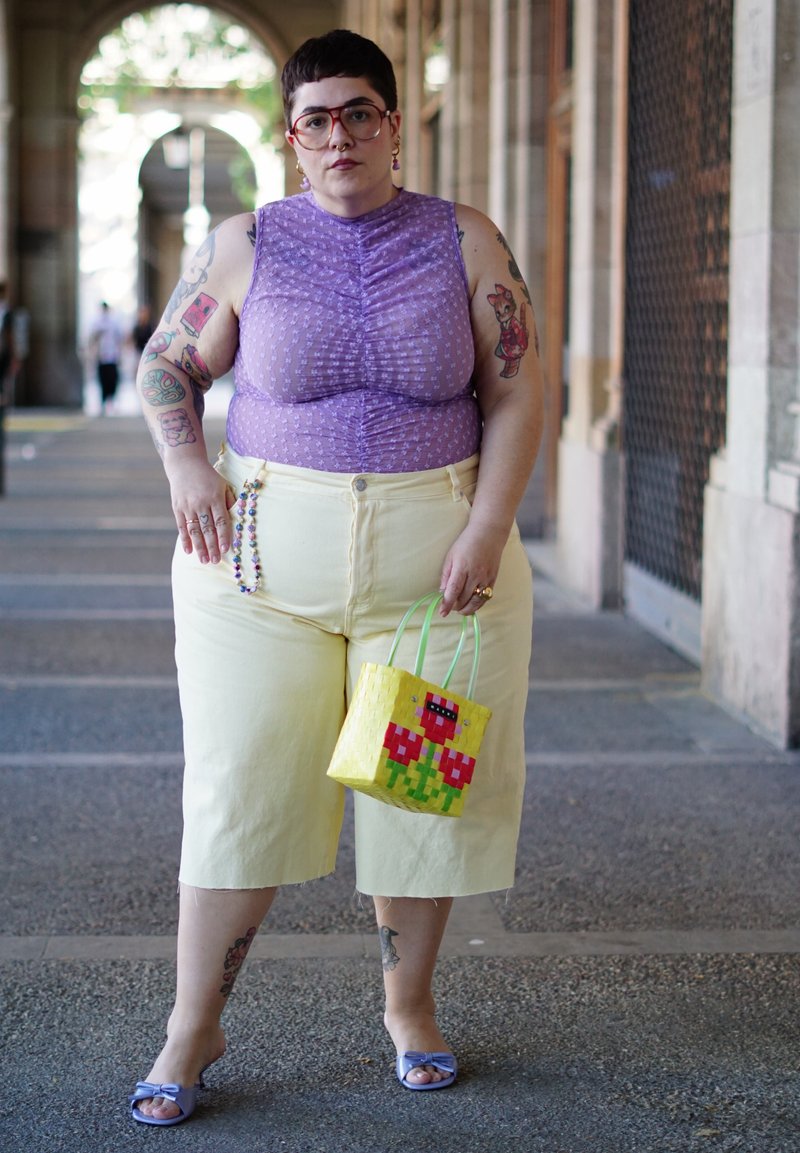 Personne aux cheveux courts et foncés avec des tatouages se tient sous une arche, portant un haut violet sans manches et un pantalon jaune pâle, tenant un sac jaune avec des fleurs.