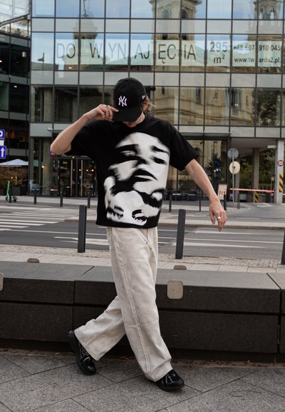 Persona con gorra negra y camiseta negra de gran tamaño con rostros abstractos, caminando por la acera de la ciudad frente a un edificio de cristal.